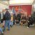 Visitors at an automotive seat exhibition, seated in large car seats on display under a red banner featuring off-road imagery.