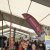 Crowded indoor trade show under a white tent with tall vertical banners and a large red flag reading 'AUTO...' (text partially visible) orients visitors down a booth-lined aisle.